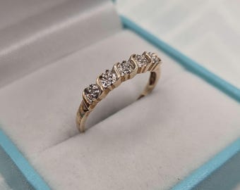 Puede incluir: Un anillo de oro con cinco piedras claras de talla redonda dispuestas en fila. La banda de metal tiene un acabado pulido y las piedras están sujetas por engastes individuales. El anillo está colocado en posición vertical en un joyero azul claro.