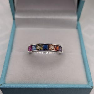 May include: A silver band ring featuring seven square-cut gemstones in a channel setting. The stones display a gradient of colours including pink, light blue, olive green, dark blue, peach, orange, and purple, set within a polished metal band.