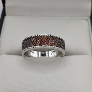 May include: A silver ring featuring a band of small, dark red gemstones. The ring has a beaded border on the top and bottom edges. The ring is presented in a grey jewellery box.