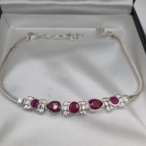 May include: A silver bracelet with a chain design, featuring six oval and teardrop-shaped red gemstones. The bracelet is presented in a black jewellery box with a white satin lining.