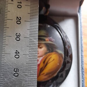 May include: A black and white cameo with a painted portrait of a person wearing a yellow jacket and a hat. The cameo is approximately 5 cm in diameter.