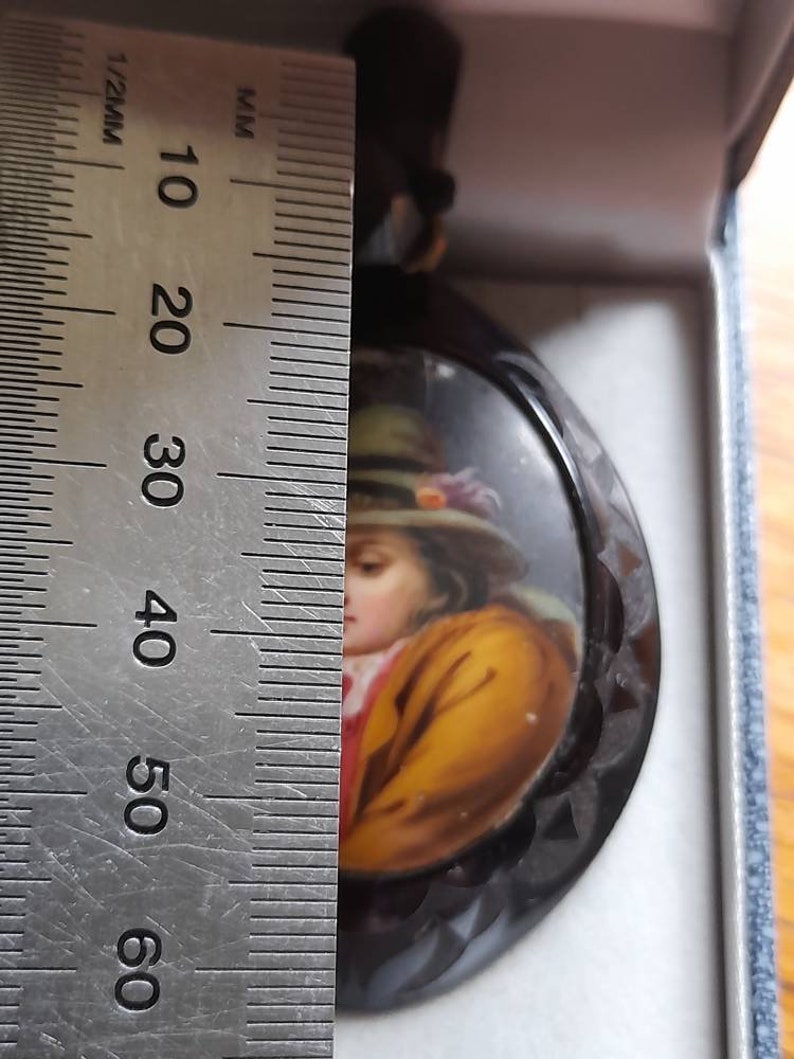 May include: A black and white cameo with a painted portrait of a person wearing a yellow jacket and a hat. The cameo is approximately 5 cm in diameter.