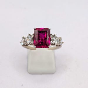 May include: A silver ring with a large, rectangular, deep pink gemstone at its centre. Either side of the main stone are smaller, clear, triangular-cut stones. The ring is displayed against a white backdrop.