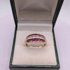 May include: A gold band ring featuring three horizontal rows of gemstones. The top and bottom rows consist of round, deep pink rubies, while the center row contains a series of clear, rectangular baguette-cut diamonds set in a channel.