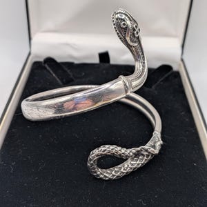 May include: A silver snake bracelet with a detailed head and tail. The bracelet is presented in a black velvet-lined box with a white interior. The snake's head is raised, and the tail is coiled, highlighting the intricate design.