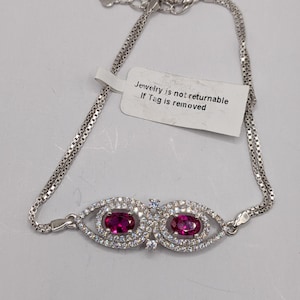 Puede incluir: Una pulsera de plata con dos gemas ovaladas de color rubí, cada una rodeada de pequeños cristales transparentes. La pulsera tiene una cadena delicada y un cierre. Una etiqueta dice "Jewelry is not returnable If Tag is removed."