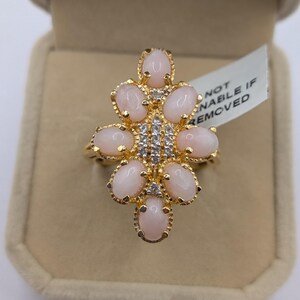 May include: A gold ring with a floral design. It features oval, pale pink gemstones set around a cluster of small, clear stones. The tag reads "NOT REMOVABLE IF REMOVED". The ring is presented in a beige box.