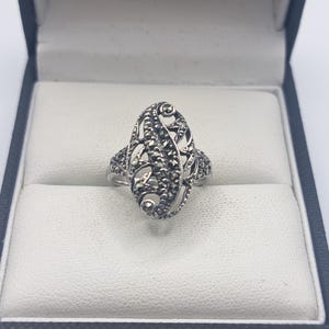 May include: A silver ring with an oval filigree design, set with small, dark stones. The ring is displayed in a black jewelry box with a white interior, showcasing its intricate details and craftsmanship.