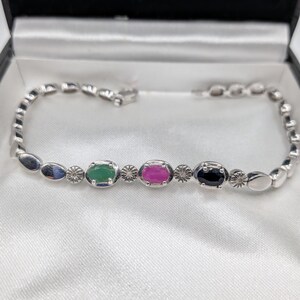 Pode incluir: Uma pulseira de prata com pedras preciosas ovais verdes, rosa e pretas, intercaladas com elementos decorativos. A pulseira é apresentada numa caixa de joalharia preta com forro de cetim branco.