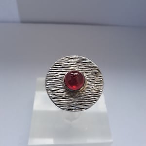 Op de afbeelding: Een zilveren ring met een getextureerde ovale band en een rode edelsteen in het midden.