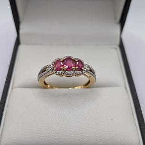May include: A gold ring with three oval-cut, vibrant pink gemstones set in the center. The ring is accented with smaller, clear gemstones and sits in a black jewelry box with a white interior.