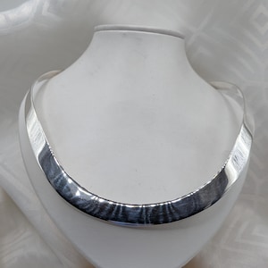 May include: A polished silver-tone metal collar necklace with a wide, curved, and rigid band design. The surface of the necklace features a subtle brushed texture that catches the light, creating a reflective finish across the entire front arc.