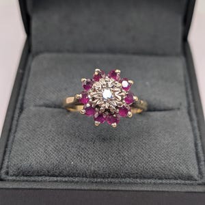 May include: A gold ring featuring a central clear gemstone surrounded by a cluster of small, round, deep red gemstones. The ring is presented in a dark grey jewellery box.