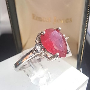 May include: A silver ring featuring a large, oval-cut, deep red gemstone. The ring has a prong setting and small accent stones. The ring is displayed in a green and gold box with the text "Ernest Jones".