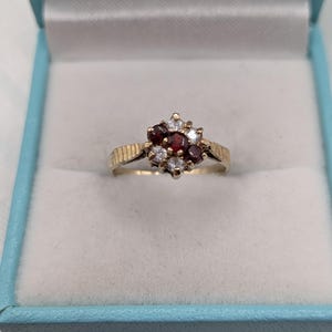 May include: A gold ring featuring a cluster setting with three deep red round-cut stones and four clear white round-cut stones arranged in a floral pattern. The band has a textured, ribbed design on the shoulders leading to a smooth gold shank.
