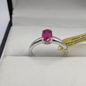 May include: A silver ring with a vibrant oval-cut ruby gemstone. The ring is set in a simple, elegant band, and is displayed in a white jewelry box. The ruby is a deep, rich red color.