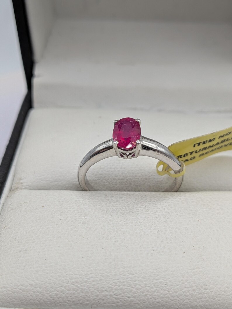 May include: A silver ring with a vibrant oval-cut ruby gemstone. The ring is set in a simple, elegant band, and is displayed in a white jewelry box. The ruby is a deep, rich red color.