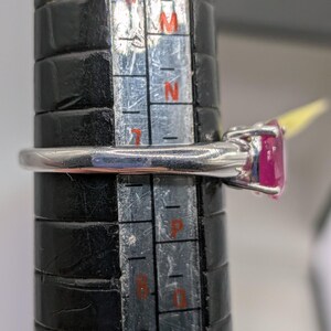 May include: A silver ring with a large, oval-cut, bright pink gemstone. The ring is being measured on a black ring sizer. The ring is a size N.