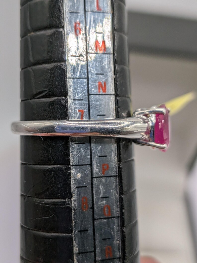 May include: A silver ring with a large, oval-cut, bright pink gemstone. The ring is being measured on a black ring sizer. The ring is a size N.