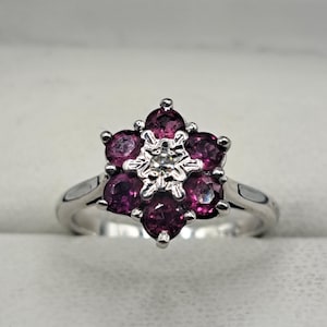 May include: A silver ring with a floral design. The flower is made up of six round, deep pink gemstones encircling a central, smaller, clear gemstone. The ring band is smooth and polished.