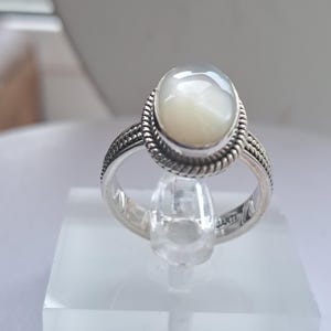May include: A silver ring with a white oval-shaped gemstone. The ring has a braided design around the gemstone.