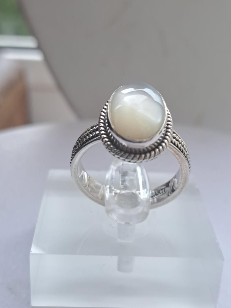May include: A silver ring with a white oval-shaped gemstone. The ring has a braided design around the gemstone.