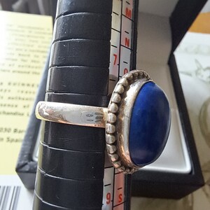 May include: A silver ring with a dark blue oval gemstone. The ring is on a sizing tool and the size is 7.