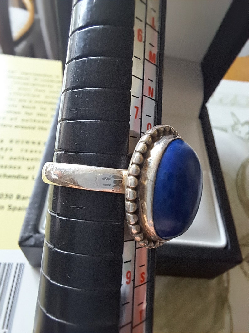 May include: A silver ring with a dark blue oval gemstone. The ring is on a sizing tool and the size is 7.