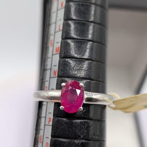 May include: A silver ring with a large, oval-cut ruby gemstone. The bright red ruby is the focal point, set in a simple, elegant band. The ring is on a black ring sizer with measurement markings.