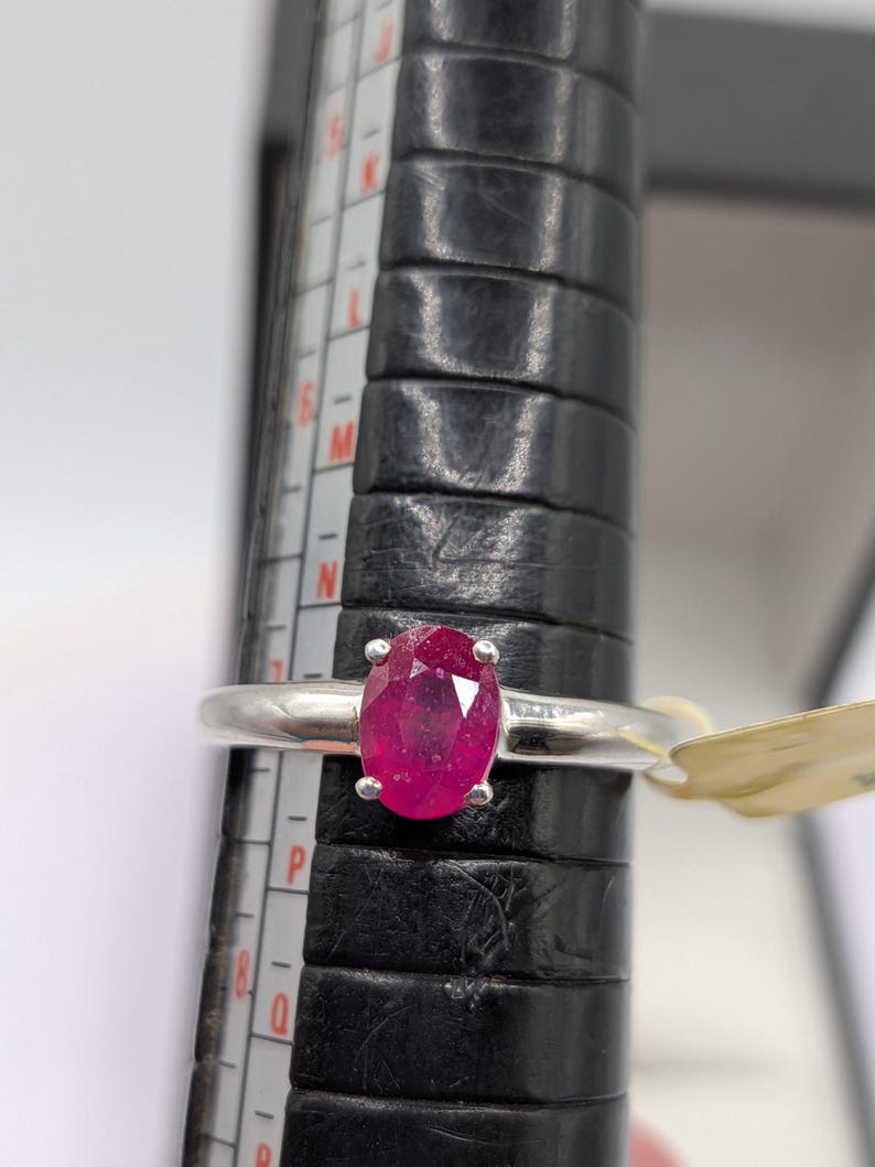 May include: A silver ring with a large, oval-cut ruby gemstone. The bright red ruby is the focal point, set in a simple, elegant band. The ring is on a black ring sizer with measurement markings.