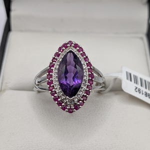 May include: A silver ring featuring a large, marquise-cut purple gemstone, surrounded by a halo of small, round red gemstones. A second halo of small, clear gemstones borders the purple stone. The ring has a split shank design.
