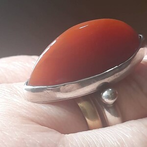 May include: A silver ring with a large, red carnelian stone set in a bezel setting. The ring is worn on a finger.