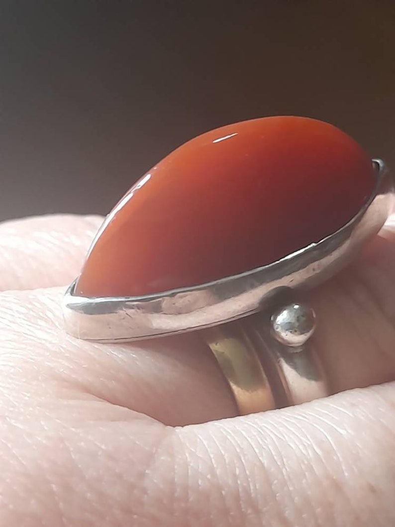 May include: A silver ring with a large, red carnelian stone set in a bezel setting. The ring is worn on a finger.