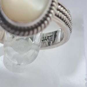 May include: A silver ring with a white oval-shaped stone set in a rope-like design. The ring is marked "ARTI" on the inside band.