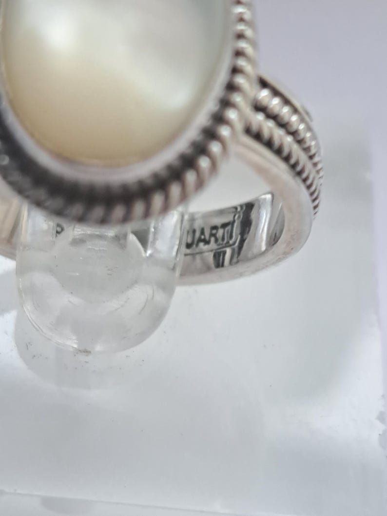 May include: A silver ring with a white oval-shaped stone set in a rope-like design. The ring is marked "ARTI" on the inside band.
