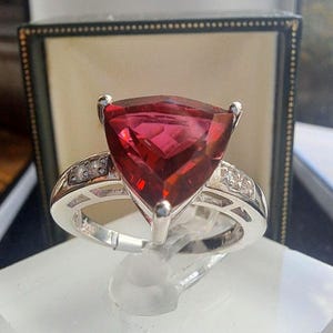 Op de afbeelding: Een zilveren ring met een grote, driehoekige, gefacetteerde, dieprode edelsteen. De band van de ring is versierd met kleinere, heldere edelstenen. De ring wordt gepresenteerd op een transparante standaard, met een donkergroene en zwarte doos op de achtergrond.