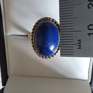 May include: A silver ring with a blue oval gemstone set in a beaded bezel. The ring is displayed in a white ring box next to a ruler.