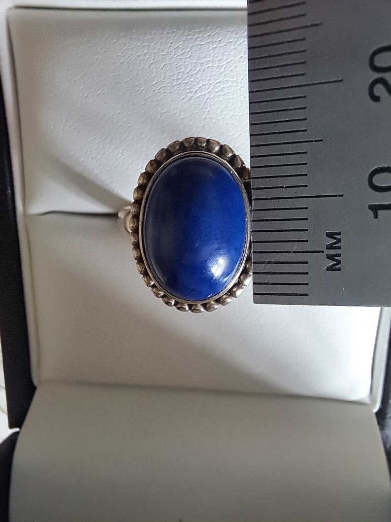 May include: A silver ring with a blue oval gemstone set in a beaded bezel. The ring is displayed in a white ring box next to a ruler.