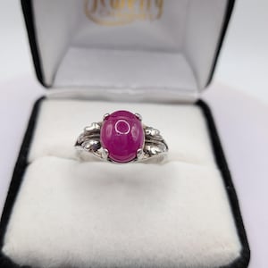 May include: A silver ring featuring a central oval cabochon-cut magenta gemstone. The metal band is detailed with leaf-like motifs on both sides of the stone. The ring is presented inside a black jewellery box with a white interior lining.