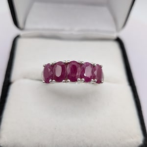 May include: A silver-toned ring featuring five oval-cut, deep pinkish-red rubies arranged in a horizontal row. Each gemstone is secured by small metal prongs, creating a continuous line of stones across the top of the band, presented in a box.