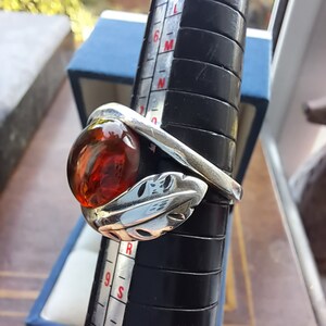 May include: A silver ring with a large, round, amber stone set in a leaf design. The ring is sized on a ring sizer.