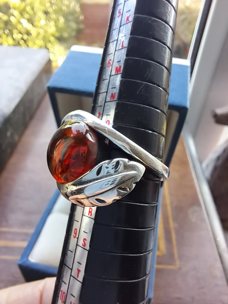 May include: A silver ring with a large, round, amber stone set in a leaf design. The ring is sized on a ring sizer.