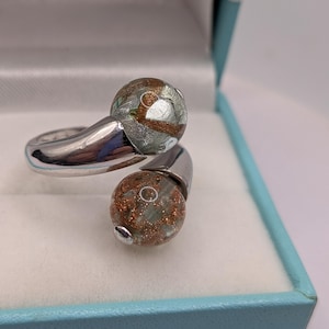 May include: A bypass ring featuring a polished silver-tone metal band that terminates in two spherical glass beads. Each bead is clear with embedded copper-colored glitter and silver-toned flecks, creating a textured, shimmering appearance.