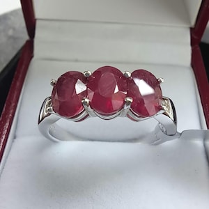 May include: A silver ring featuring three oval-cut, deep red rubies. The rubies are set in a row, with small accent stones on either side. The ring is displayed in a red and white jewelry box.
