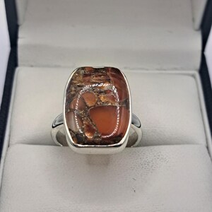 May include: A silver ring with a large, rectangular copper turquoise stone. The stone has a mix of brown, orange, and gold tones. The ring is set in a simple, polished silver band, and is displayed in a jewellery box.