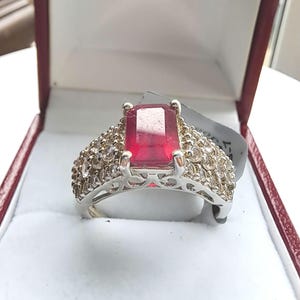 Puede incluir: Anillo de plata con una gran gema roja rectangular. La banda está adornada con gemas claras más pequeñas. El anillo se presenta en una caja de joyería roja con interior blanco, ideal para regalo o exhibición.