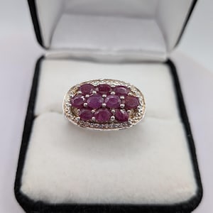 Peut inclure: Une bague argentée présentant un groupe ovale de neuf pierres précieuses rose-rouge foncé taillées en ovale. Le groupe central est entouré d'une bordure de petites pierres claires et scintillantes serties dans un cadre métallique texturé.