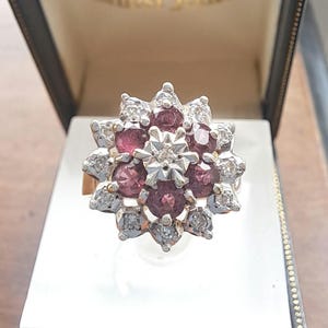 Puede incluir: Un anillo dorado con un diseño floral. El centro del anillo presenta un grupo de piedras preciosas de color rojo oscuro, rodeadas de piedras más pequeñas y transparentes. El anillo se muestra en una caja de joyería negra con interior blanco.