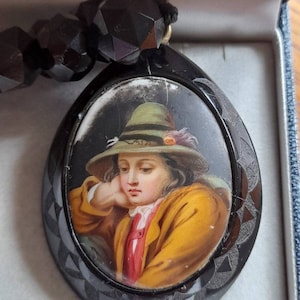 May include: A black jet necklace with a cameo pendant featuring a portrait of a young person wearing a green hat. The cameo is set in a black oval frame with a decorative design.