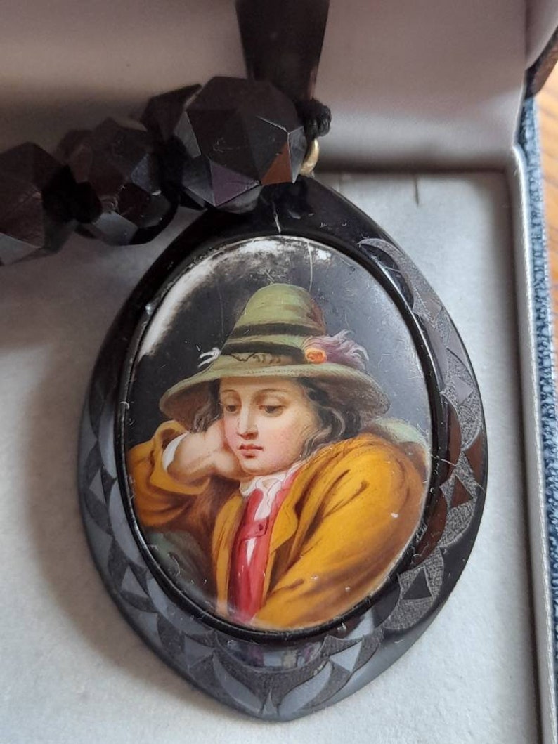 May include: A black jet necklace with a cameo pendant featuring a portrait of a young person wearing a green hat. The cameo is set in a black oval frame with a decorative design.